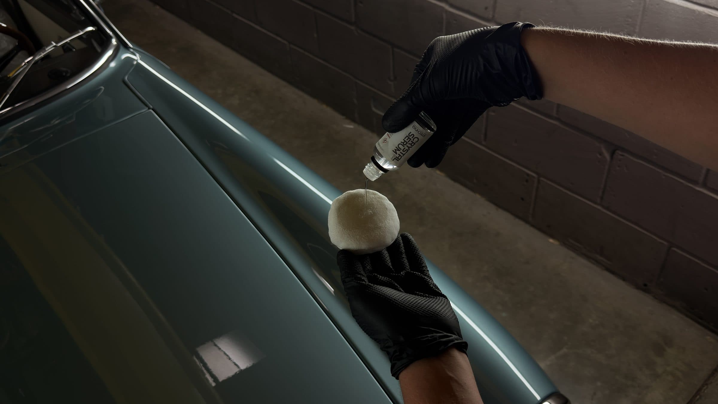 Gloved hands loading a coating applicator beside the bonnet of a light blue MGB GT