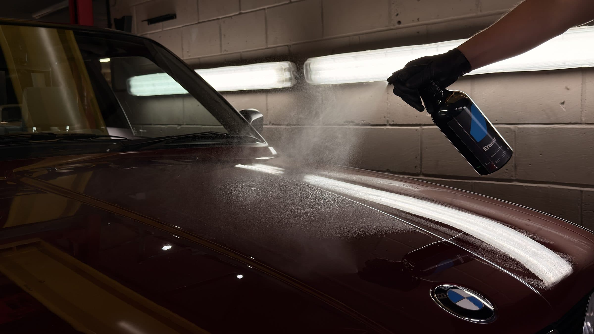 Gloved hand misting panel prep from a spray bottle onto a dark red BMW bonnet