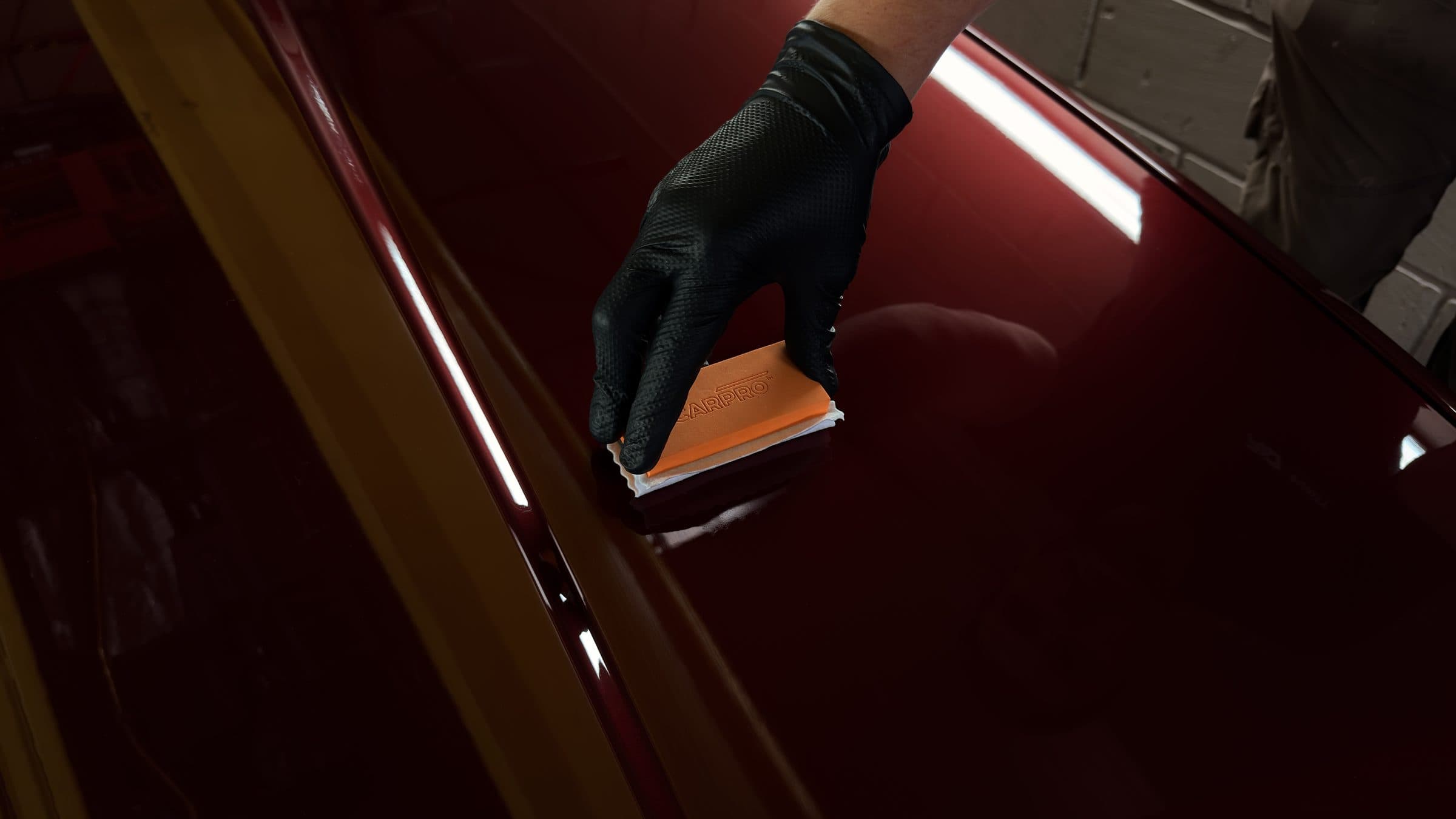Black-gloved hand drawing an orange coating applicator across the dark red paint of a car