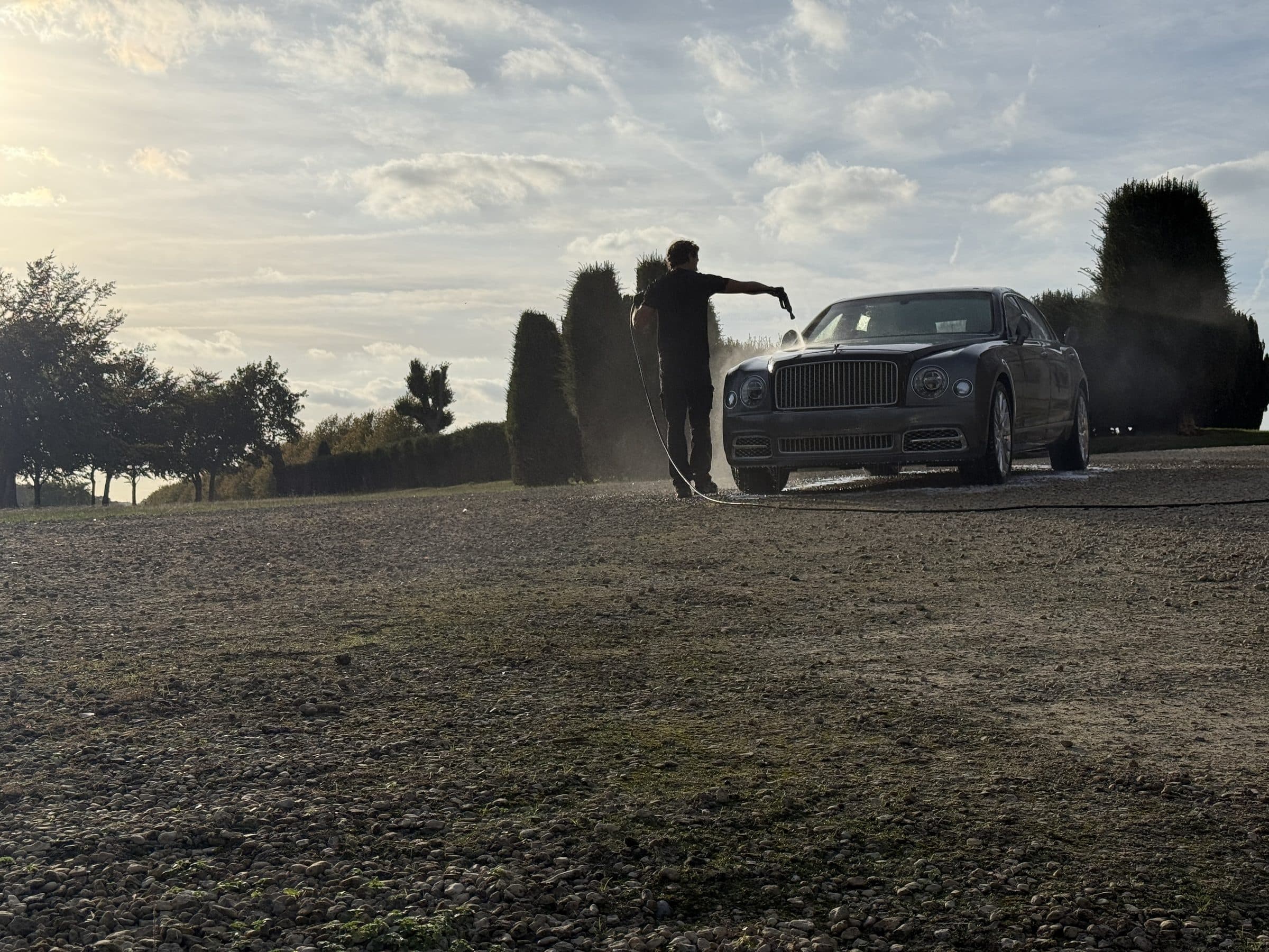 Detailer rinsing a Bentley Mulsanne on a gravel drive, backlit by a low sun