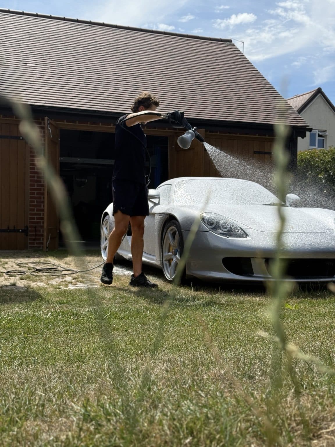 Detailer rinsing a silver Porsche Carrera GT beside a timber garage